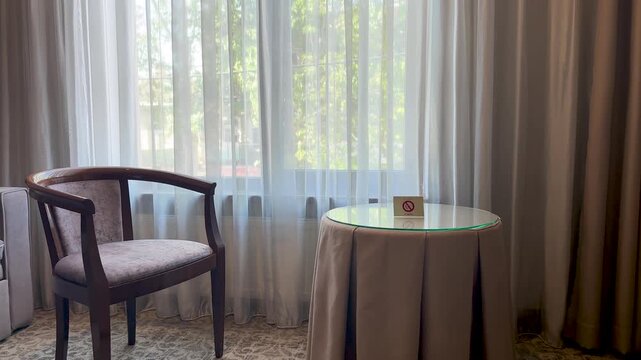 Interior of a cozy hotel room with a wooden armchair, a round glass table with a no smoking sign, and sunlight filtering through sheer white curtains