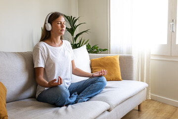 Fototapeta premium Young woman meditating on the couch listening to guided meditation using wireless headphones. Copy space.