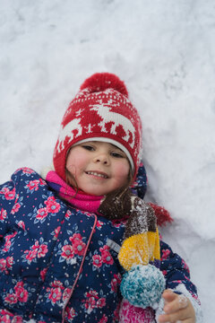 Portrait of smiling girl in red hat lying in snow holding mitten