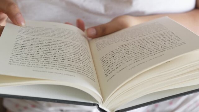 Close-up of a woman's hand gently pressing and smoothing an open book page while reading, with clearly visible printed text and soft natural window light illuminating.