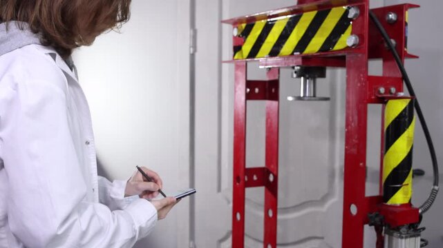 Engineer in lab coat taking digital notes on smartphone near industrial hydraulic press