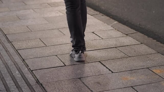 Night sidewalk closeup of footsteps, panning lowerleg shot with sneakers, tactile paving and wet reflections from streetlight, quiet evening commute with cinematic rhythm and slow steady pace