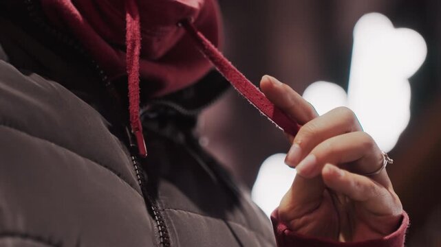 Nighttime hands adjusting. Closeup of winter urban scene. Intimate gesture of adjusting hood in city at night. Close view of hands pulling hoodie string amidst city lights and chilly atmosphere
