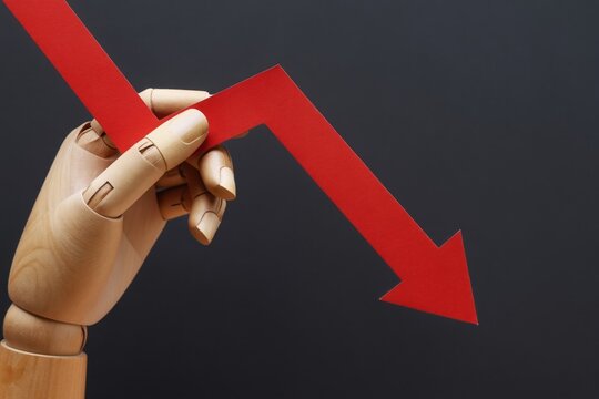 Wooden hand holds a red arrow pointing down. This symbolizes financial decline, cost reduction, and falling prices. Concept of economic recession and lower living standards.