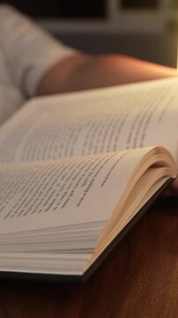 A woman's hand turns pages of an open book resting on a dark wooden table, bathed in warm golden lamp light, creating a cozy intimate reading atmosphere with soft bokeh in the background.