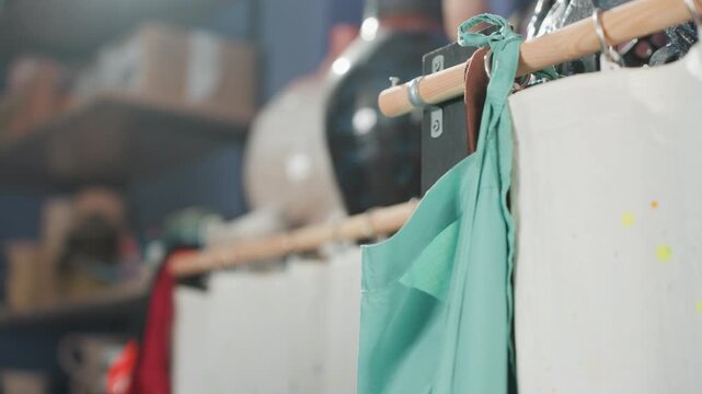 Apron tied on studio rod, pottery shelf visible white apprentice secures teal apron against paintsplattered curtain in cozy workshop, hands focused on neat ties and hanging display