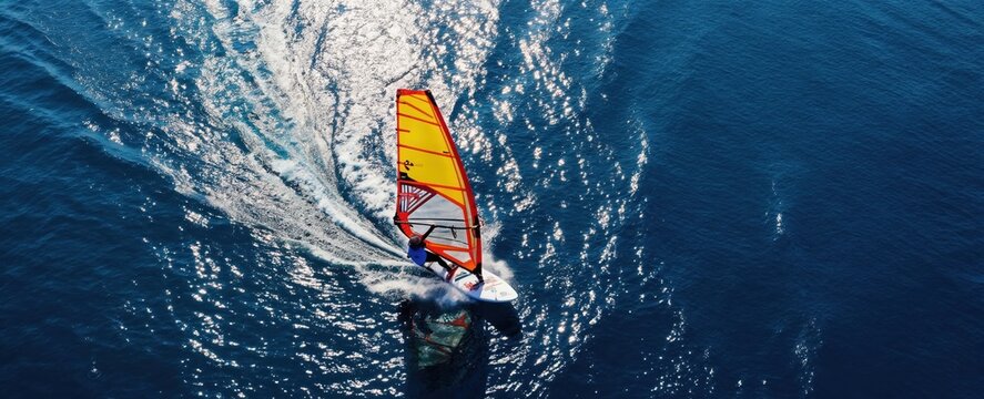 Windsurfer rides fast across deep blue ocean water creating white splash waves. Yellow sail catches wind power. Drone view from high above shows surfer on surfboard moving at speed.