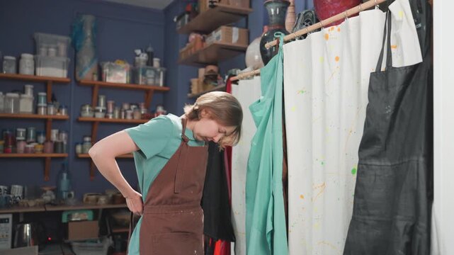 White ceramicist organizing aprons in studio with shelves of glazes and jars. Hands adjust straps, check supplies, tidy storage and prepare workspace confident practical rhythm, small studio energy