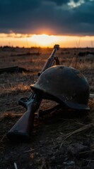 Obraz premium Old military helmet and rifle rest on dry grass field. Sunset sky with dark clouds above desolate battlefield. Quiet dusk scene offers somber mood.
