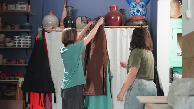 Pottery workshop apron fitting joyful teamwork, friends try on aprons amid shelves of pots, one adjusts straps while other laughs, shared excitement and playful high five, studio tools visible, ready
