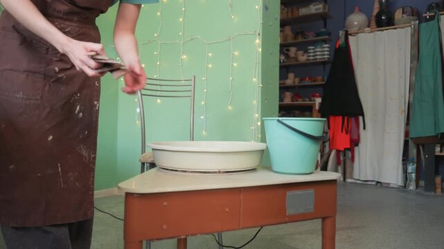 Young white woman shaping clay on wheel wearing brown apron in cozy pottery workshop with string lights and shelves, hands smoothing wet clay, bucket and sponge nearby, focused artisan lesson showing