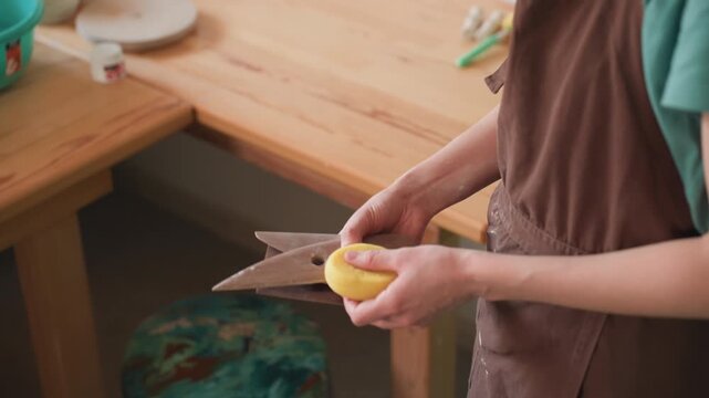 Hands prepping pottery workshop wooden table with sponge and tools apron visible, surface smoothed before wheel work measured motions and calm studio atmosphere assistant arranging bench for next