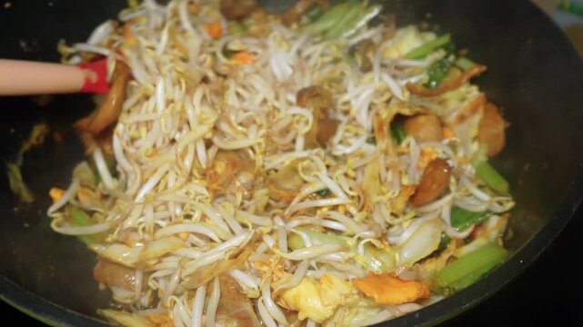 The process of cooking special homemade stir-fried flat rice noodles (Kwetiau Goreng). The flat noodles are cooked with eggs, bean sprouts, bok choy, cabbage, and sliced meatballs