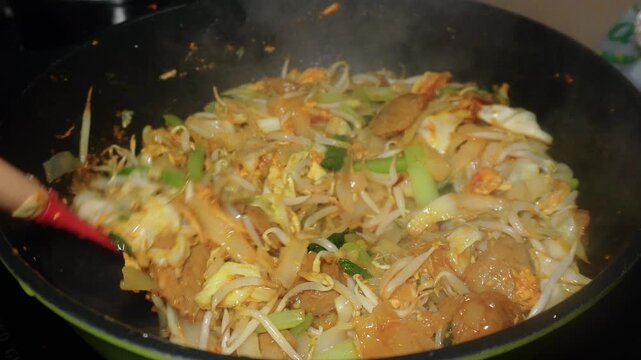 The process of cooking special homemade stir-fried flat rice noodles (Kwetiau Goreng). The flat noodles are cooked with eggs, bean sprouts, bok choy, cabbage, and sliced meatballs