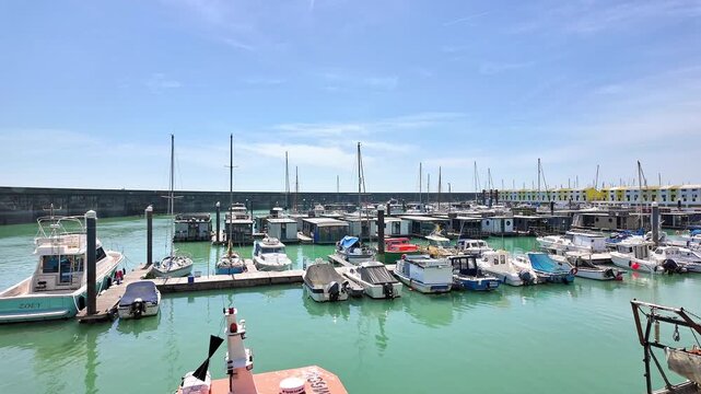 Panning shot of boats moored in Brighton Marina on a hot, sunny day, capturing gleaming hulls, calm waters, and a lively harbour atmosphere beneath clear blue skies.