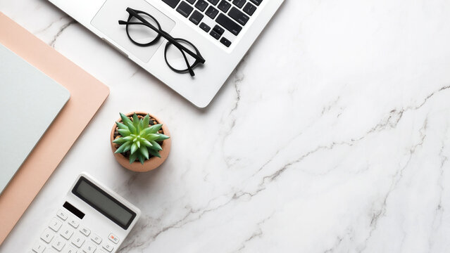 Silver laptop, glasses, tea cup on a bamboo tray with a gold spoon, notebook, succulent plant and pen on white marble desk create a productive modern workspace. Top view with copy space.