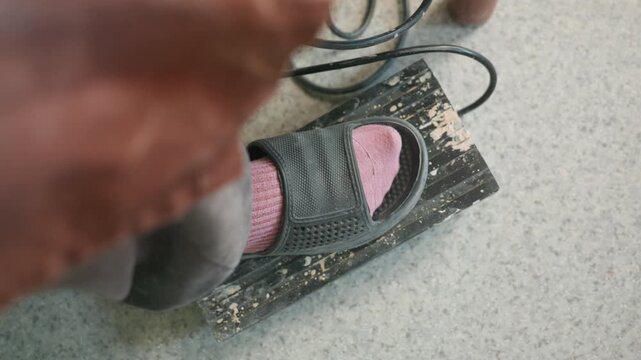 Artisan hand adjusts pottery pedal cord up close. Experienced potter tunes foot control with lace cord beside pink socked foot, weathered wooden pedal and concrete bench visible, close angle capturing