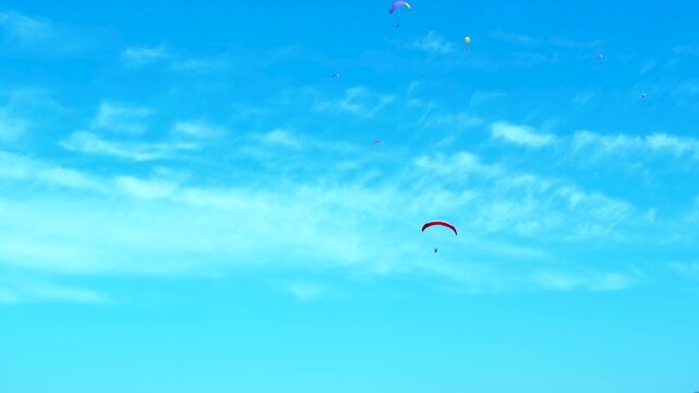 Paraglider flying in blue sky with distant colorful parachutes. Red paraglider soars through the vast blue sky, adorned with scattered white clouds, while other parachutes drift in the distance