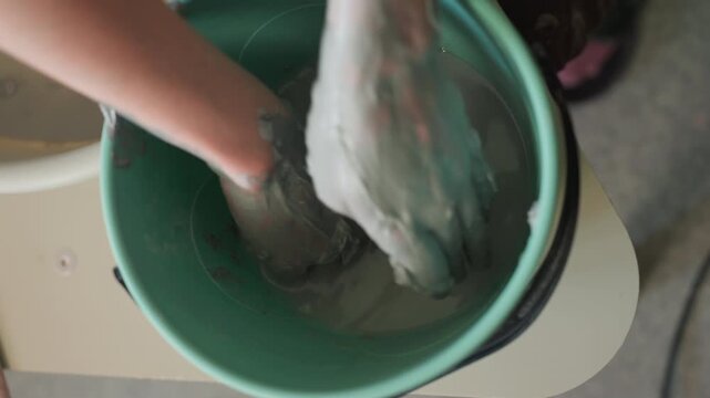 Hands mixing clay slip in workshop, green bucket of gray slip stirred and kneaded on workbench, potter hands coated in wet clay, tactile texture and gentle motion, quiet studio atmosphere highlighting