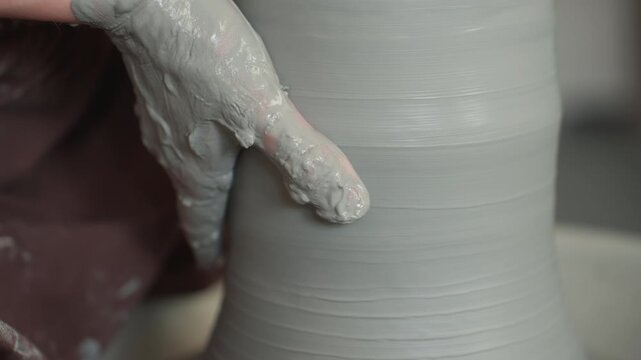 Pottery workshop caucasian potter smoothing clay cylinder, closeup hands coated in slip refining wall profile with gentle pressure, soft neutral light, contemplative tactile focus and quiet studio