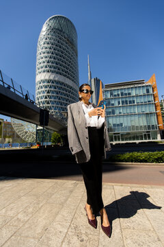 Businesswoman checks smartphone in urban setting with modern buildings and clear skies