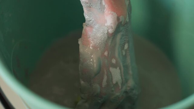 Closeup of hands stirring wet clay mixture. Detailed view of hand combining clay slip in workshop bucket. Intimate shot of hands thoroughly blending gray clay for pottery creation process