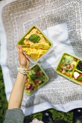 Fototapeta premium A hand holds a green container of chips and dip, surrounded by vibrant salads and snacks laid out on a white blanket in a sunny park. A perfect day for outdoor dining
