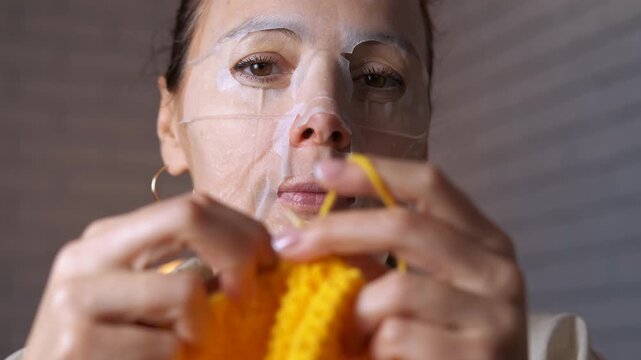 Woman with facial sheet mask knitting yellow wool. Relaxed woman wearing a cosmetic sheet mask on her face and knitting with yellow wool yarn, combining self-care beauty routine with a calming hobby