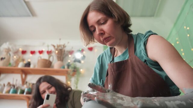 Woman shaping clay on wheel closeup. Concentrated artisan demonstration showing wet hands molding tall vessel, detailed texture and technique, shelves with jars and glazes in background, subtle studio