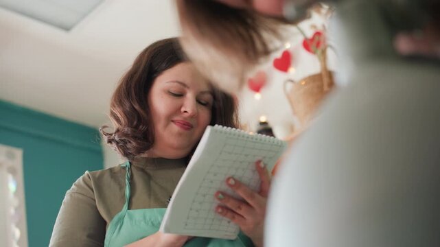 Potter planning in pottery workshop notebook jotting glaze notes and lesson plan, green apron, ceramic vase in foreground, warm ambient lighting, heart garland decor, studio shelves, focused smile,