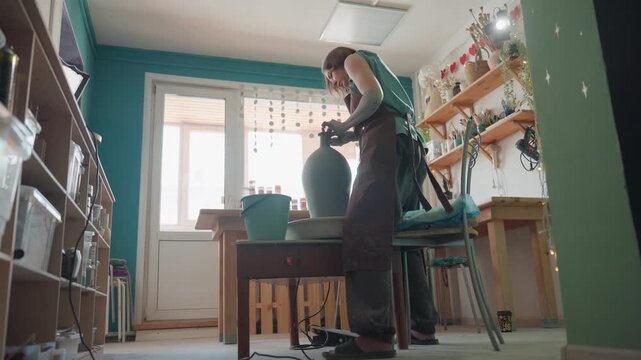 White woman finishing tall vase on wheel in bright pottery workshop, standing at workbench with hands smoothing form, shelves of ceramics and sunlight through window, calm proud mood and tactile final