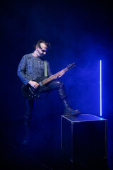 Obraz premium Adult man playing black electric guitar with foot on box in dark smoky stage with blue neon light