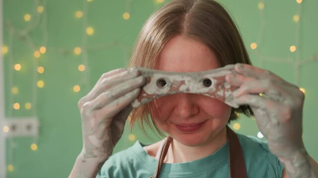 Female artist displays clay work. Caucasian woman with clay mold under playful fairy lights background. Female ceramicist in vibrant studio displaying sculpted clay piece amid whimsical lighting