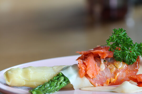 Green and white cooked asparagus wrapped in a piece of ham with cheese, a piece of salmon and parsley on a plate.