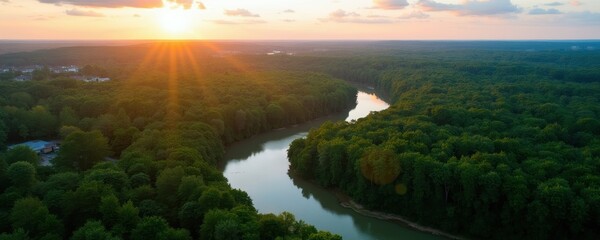 Obraz premium Aerial view of winding river through dense green forest at sunset. Sunbeams fan out over trees and sky with clouds. Distant town visible on horizon.