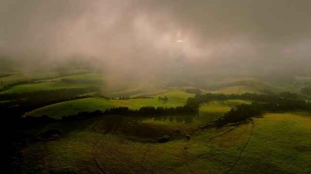 Aerial view of rolling green hills on Sao Miguel Island, bathed in soft sunset light with dramatic patches of mist and scattered clouds over the pastoral landscape.