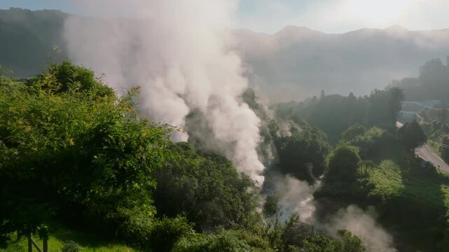 Aerial view of powerful geothermal steam vents rising from a rocky, mineral-rich ravine in Furnas, Sao Miguel Island, surrounded by lush vegetation.