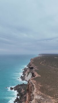 Vertical drone shot sliding laterally above the Bunda Cliffs, moving from the cliff edge out toward the hazy ocean under an overcast sky
