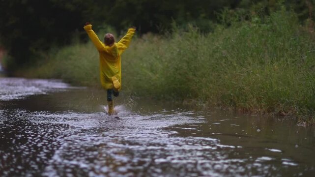 Happy An eight-year-old girl wearing yellow rubber boots and a yellow raincoat Jumping in a puddle after a summer rain. screen free kids, backyard games