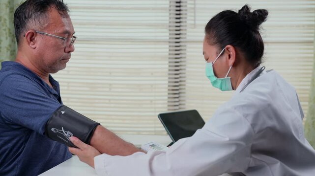 Professional Asian female doctor wearing protective mask checks spectacled senior male patient blood pressure inside bright clinic office, placing black cuff around arm, monitoring vital health.