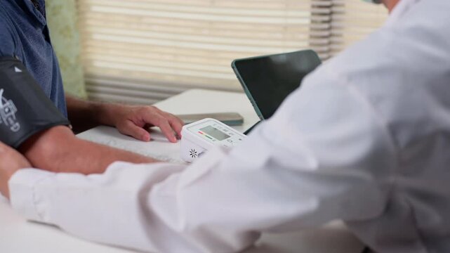 Male doctor wearing medical coat measures older patient's blood pressure inside clinic office room, operating digital monitor device atop desk near blinds, checking vital health signs very carefully.