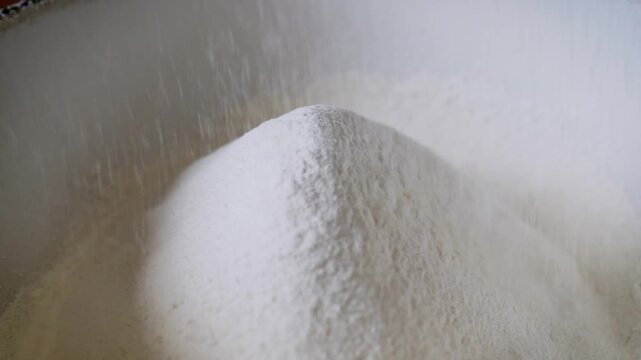 White wheat flour falls steadily onto a growing mound inside a white mixing bowl. The fine powder creates a soft texture as it is sifted during baking. Bright lighting highlights the