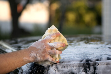 Fototapeta premium a hand with a washcloth and soap suds washes the hood of the car. Man washing car hood with washcloth and shampoo, auto covered with foam, car wash service. cleaning the black surface