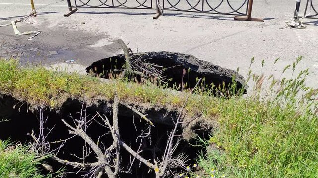 Deep hole in asphalt road with fallen branches and metal fences, sinkhole, video with zoom