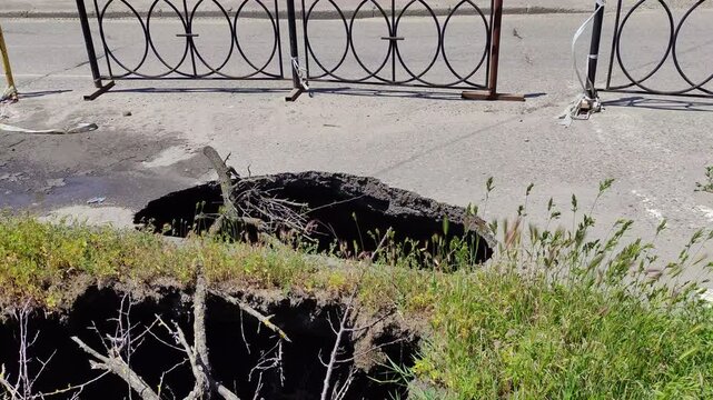 Deep hole in asphalt road with fallen branches and metal fences, sinkhole, wind weather