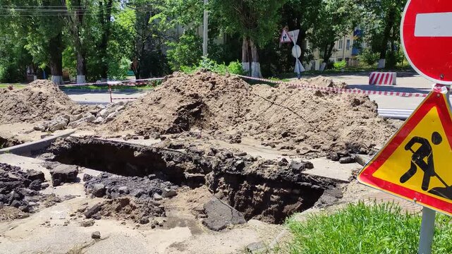 Deep trench in asphalt road with dirt mounds and red warning signs, summer day