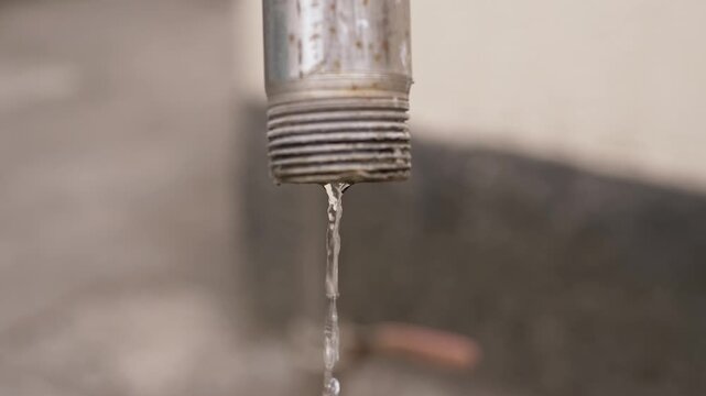 A close-up view of a thin stream of water leaking from the threaded end of a metal plumbing pipe. Concept of water scarcity, utility waste, plumbing issues, and conservation awareness.