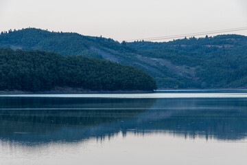Fototapeta premium Tranquil landscape of Lake Plastira in Greece featuring forested mountains softly reflecting on the calm, mirror-like water surface. Scenic natural reservoir view for travel stock media.