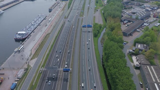 Aerial drone footage flying above the A10 motorway in Amsterdam with moving traffic, urban infrastructure, waterways and harbor facilities. Modern transportation network viewed from above.