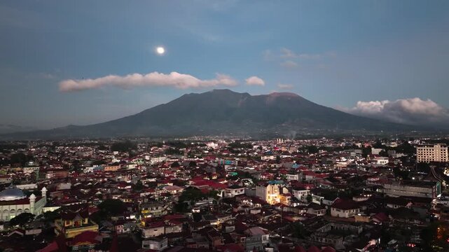 Aerial drone night view of Bukittinggi city lights glowing under a full moon beside Mount Marapi volcano, dramatic nightscape of West Sumatra Indonesia with illuminated urban skyline surrounded